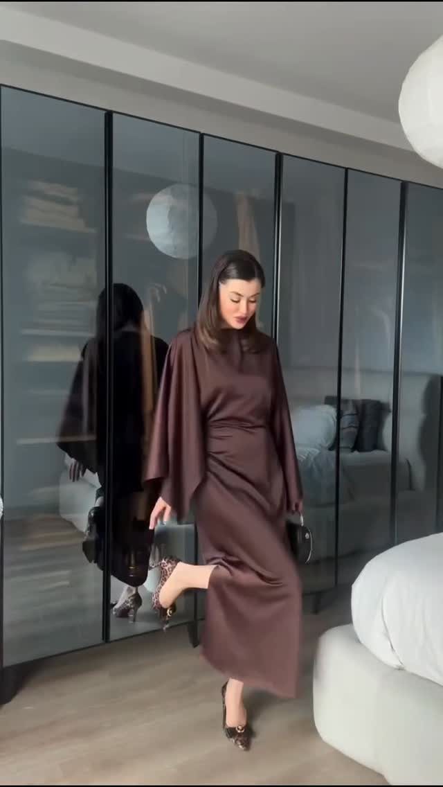 Woman wearing a long chocolate brown satin dress with flowing sleeves in a modern room.