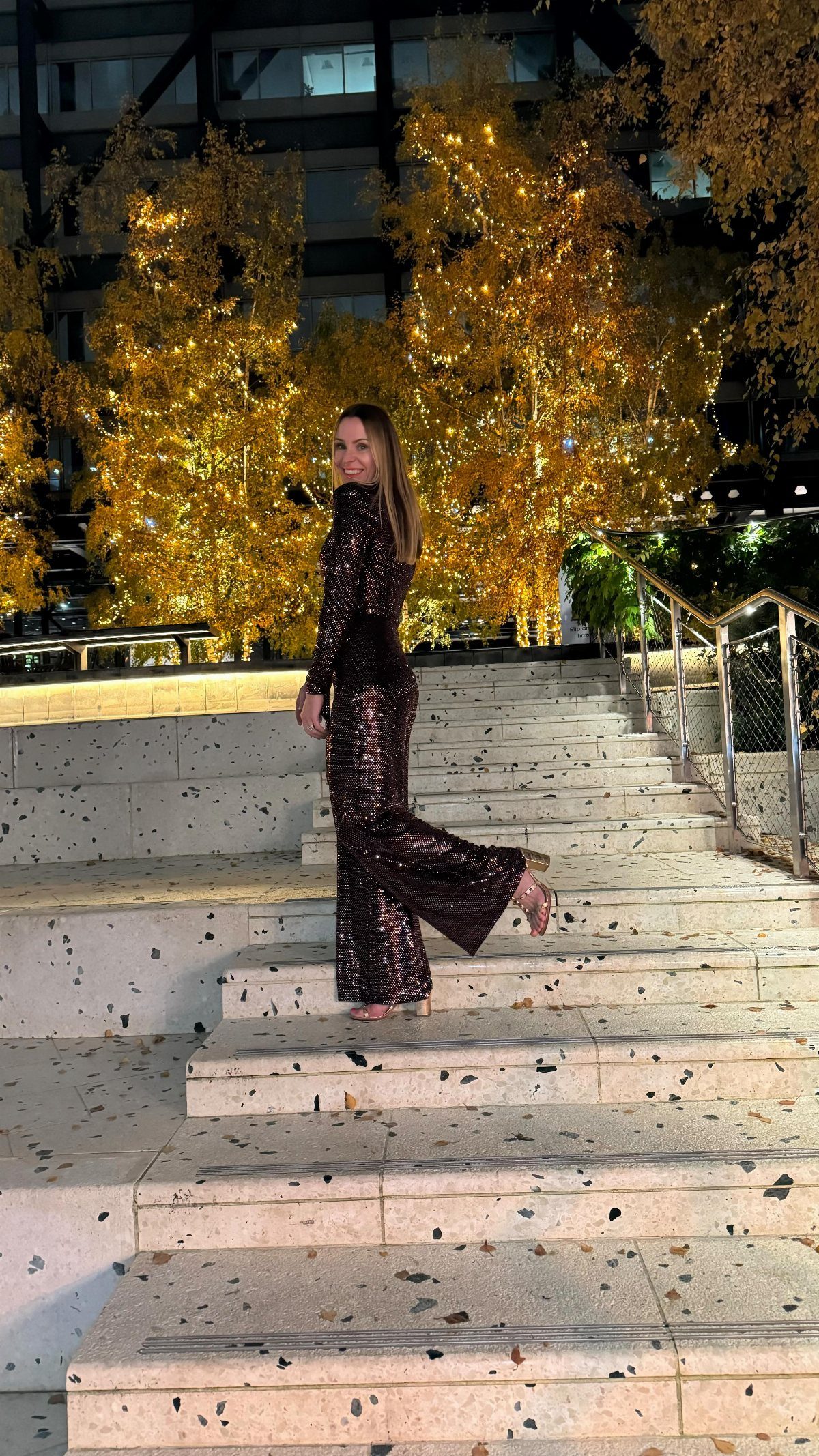 Woman in a sparkly dark jumpsuit posing on stone stairs at night.