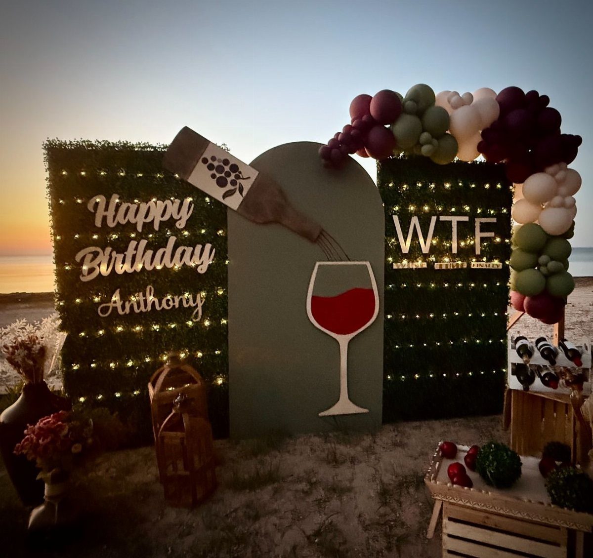 Birthday backdrop with wine decorations and balloon arches on a beach at sunset.