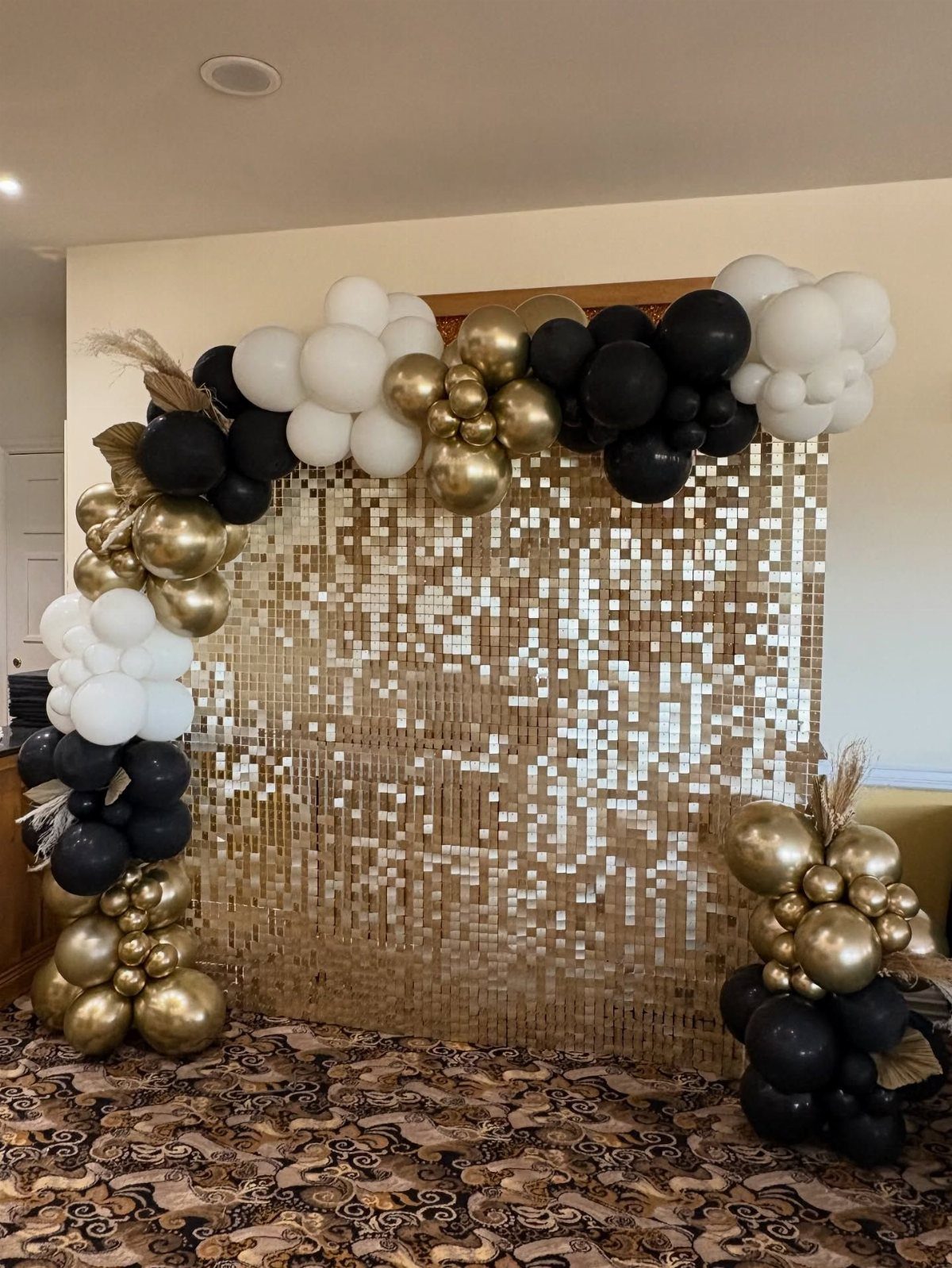 Gold sequin backdrop framed by black, white, and gold balloon clusters on a patterned carpet.