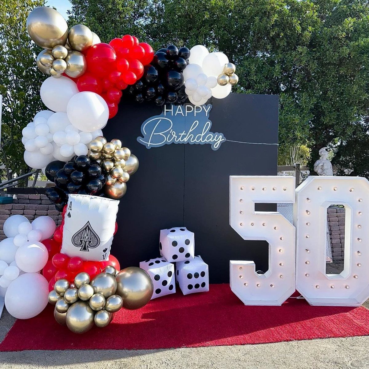 Birthday decorations with large marquee numbers fifty, dice, and a red, black, and gold balloon arch.