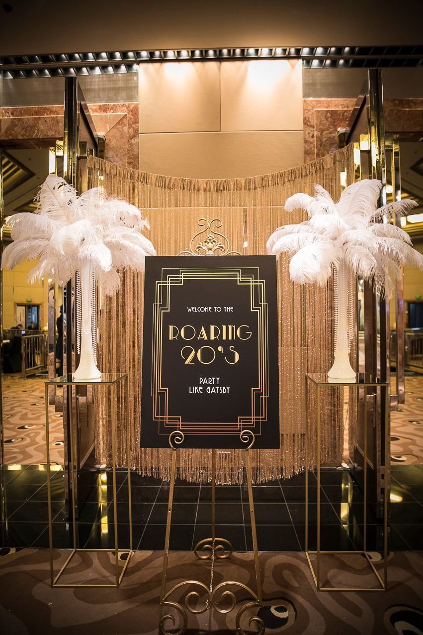 A black and gold Gatsby themed sign flanked by white feather decorations.
