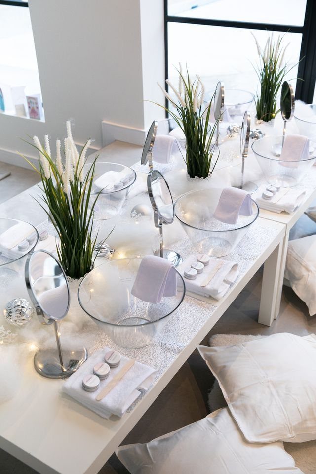 White table setup with glass bowls, mirrors, small containers, and green plants for a spa day.