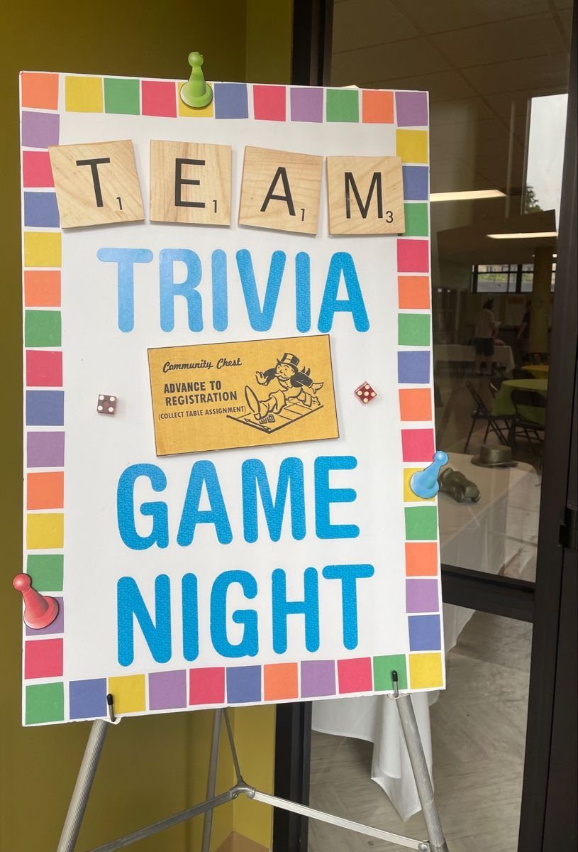 A trivia game night poster with wooden letter tiles and colorful game pieces on an easel.