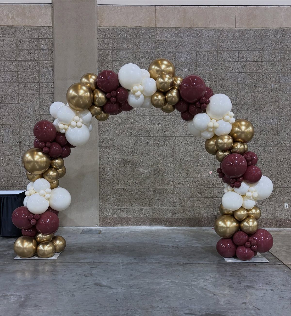 A large balloon arch featuring white, gold, and burgundy balloons against a brick wall.