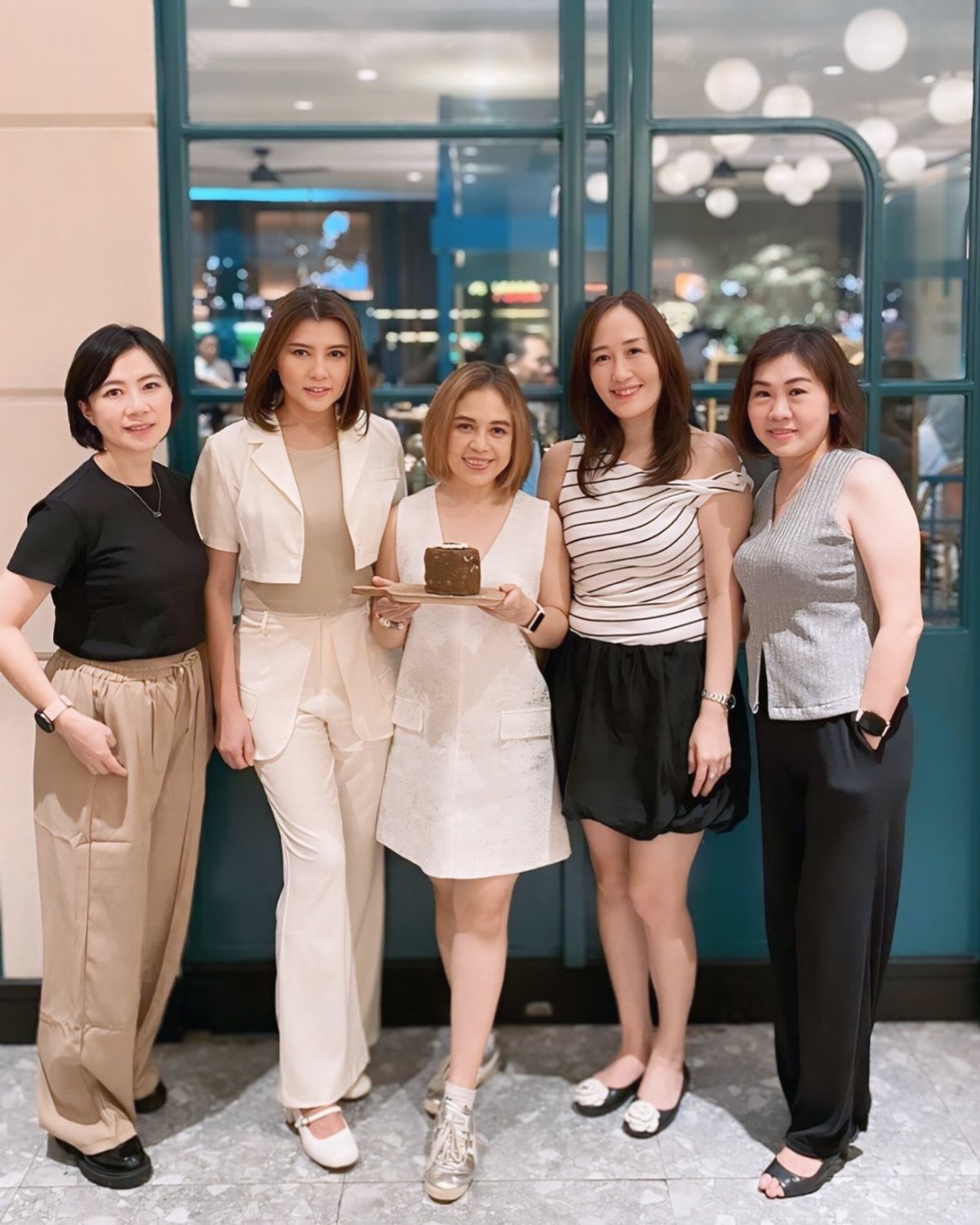 Five smiling women standing together outdoors, with one woman holding a small chocolate cake.