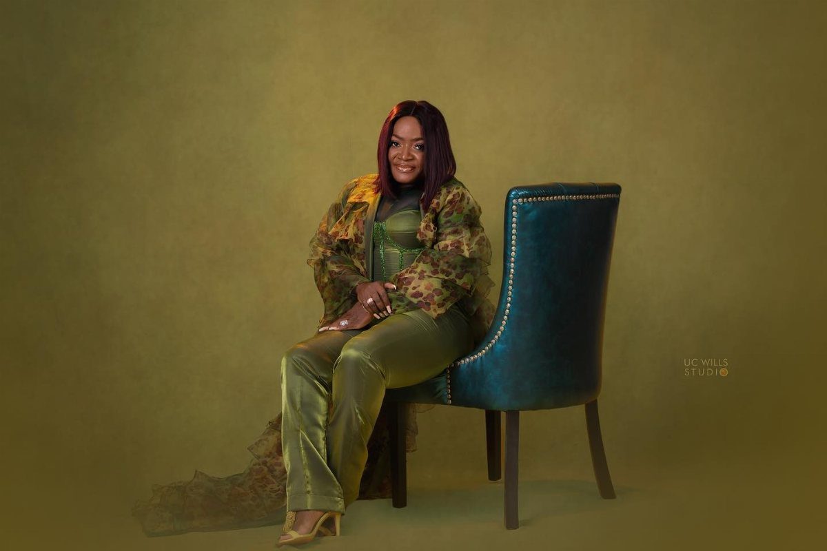 A smiling woman in green patterned clothing sitting on a teal velvet chair in a studio.