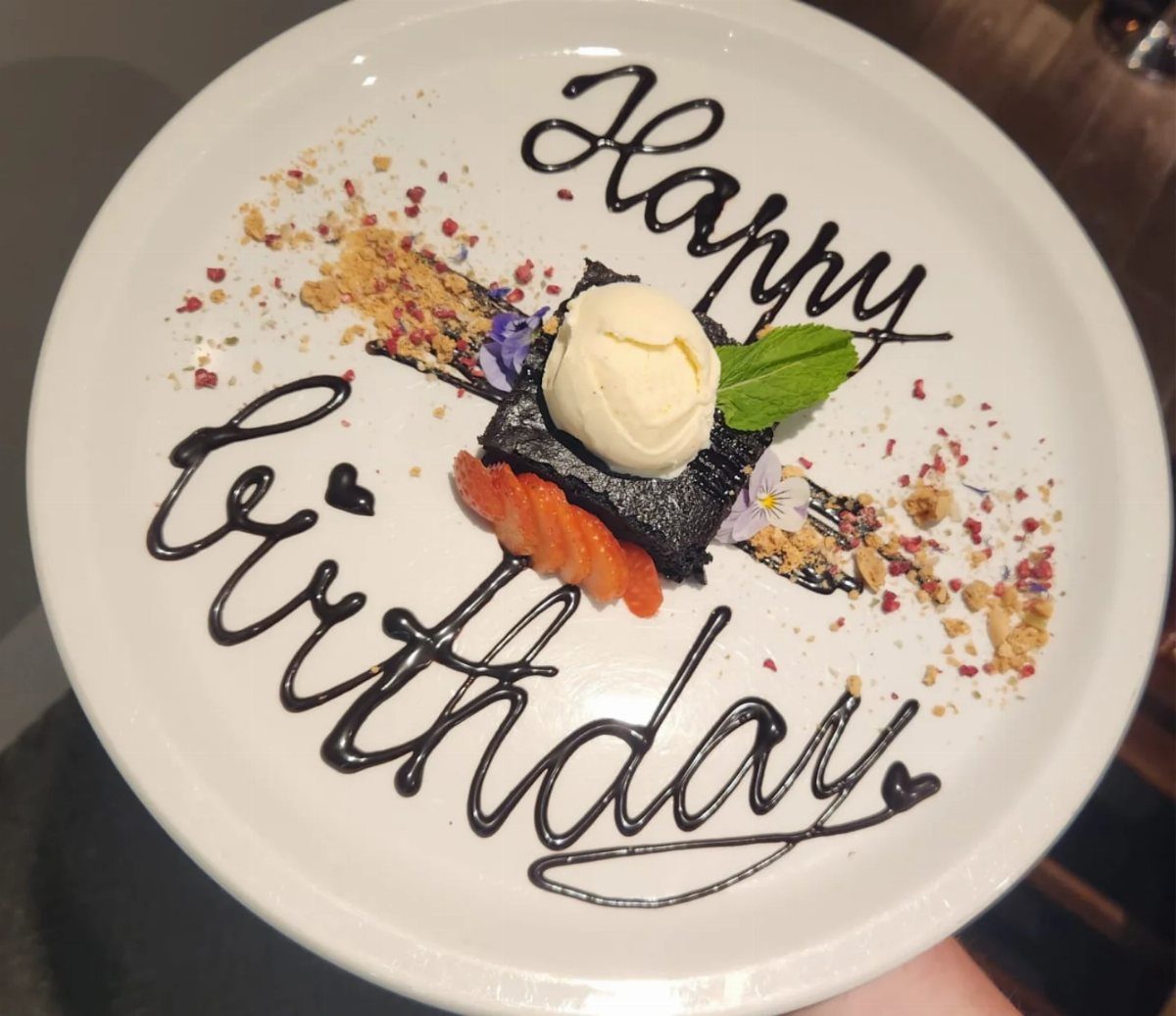Chocolate brownie with ice cream and strawberries on a plate with Happy Birthday written in chocolate.