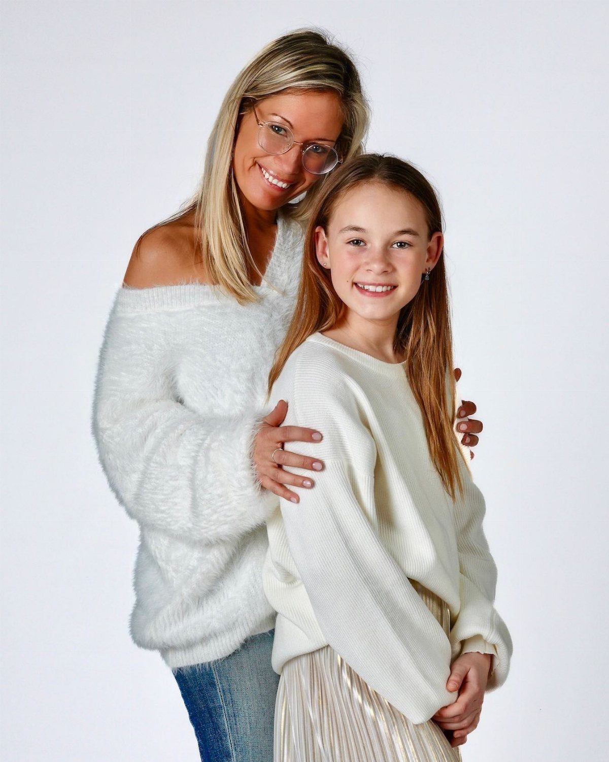 A smiling blonde woman and a young girl posing together in white sweaters against white background.