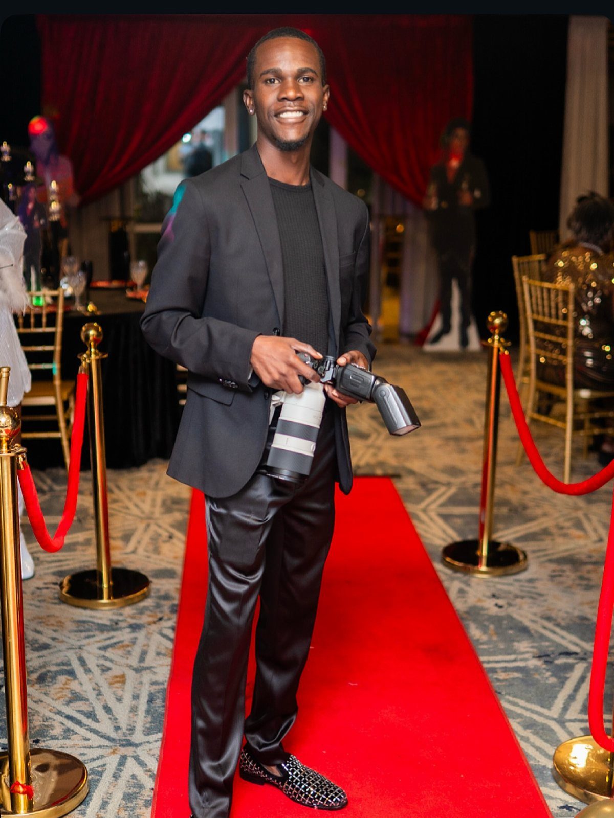 A smiling man in a black suit holding a professional camera on a red carpet.