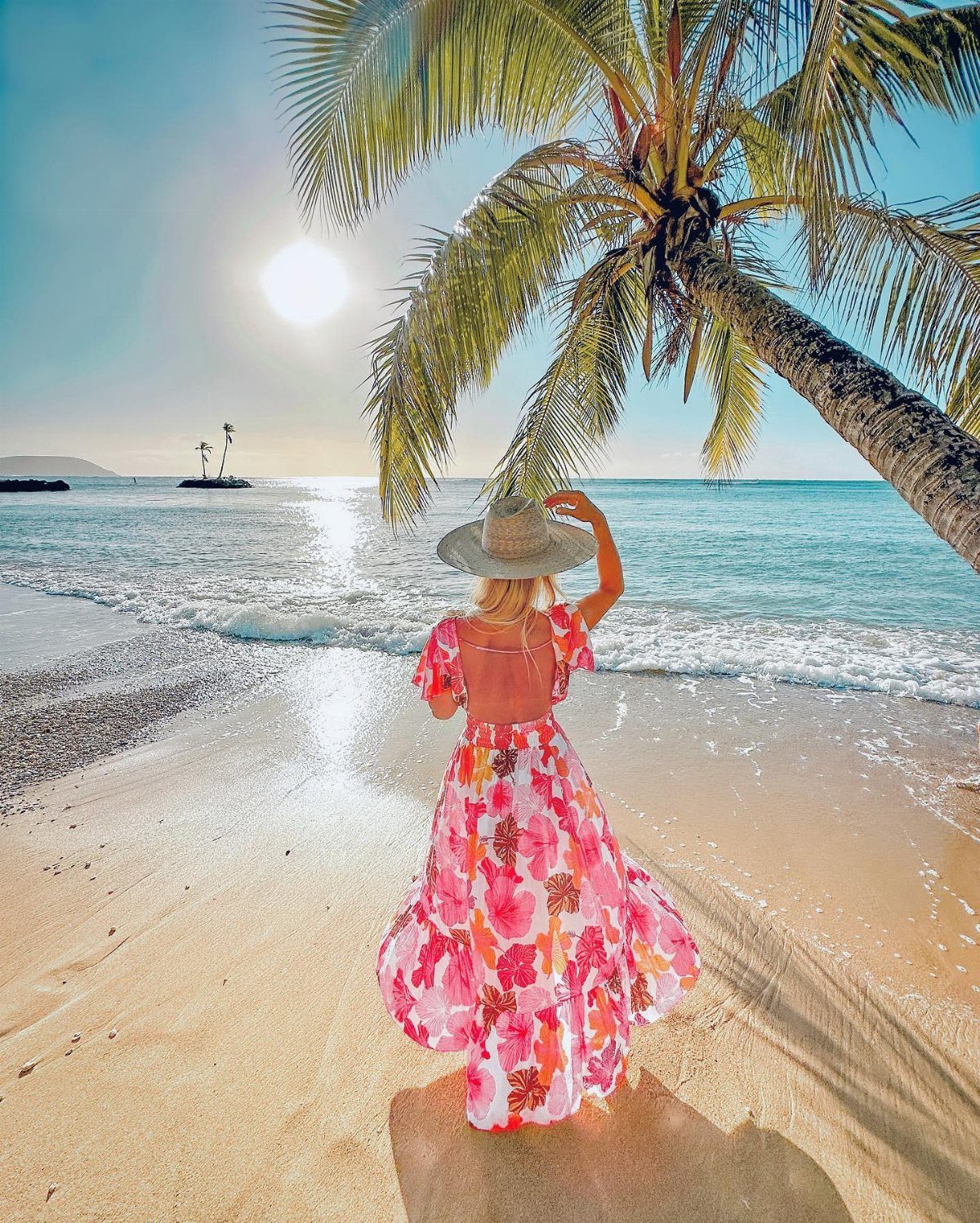 Woman in a pink floral dress and straw hat standing on a sunny tropical beach.