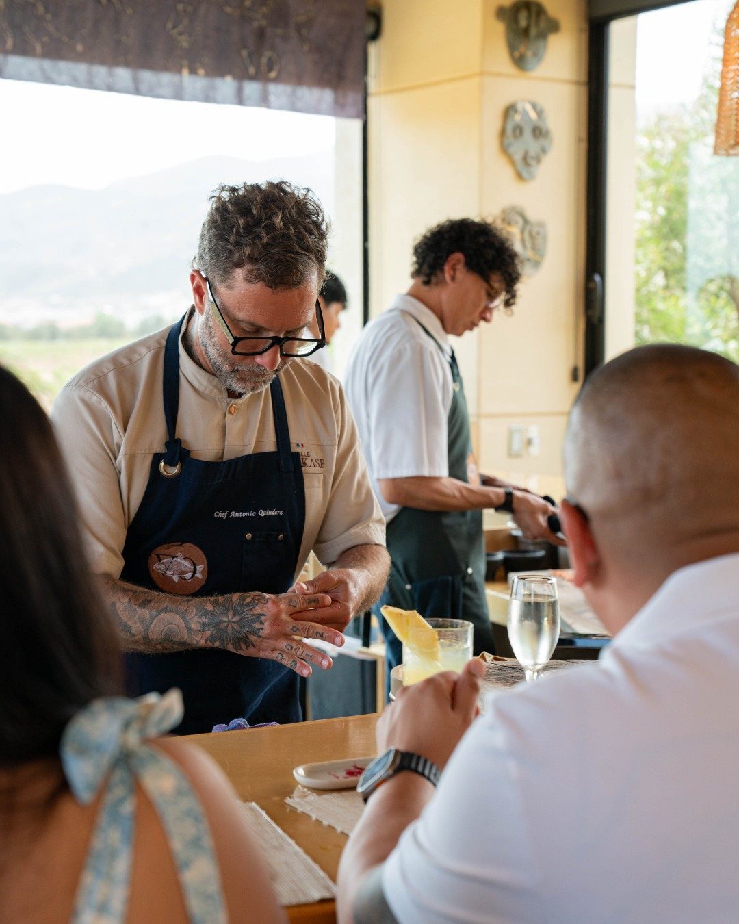Chefs preparing food at a counter for guests seated at a dining table.
