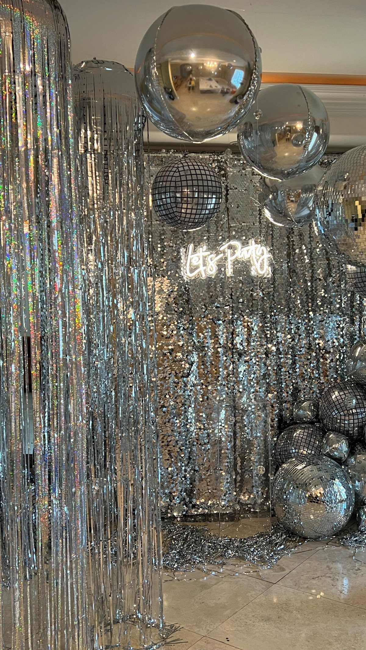 Silver disco balls, tinsel curtains, and large balloons decorating a party space with neon signage.