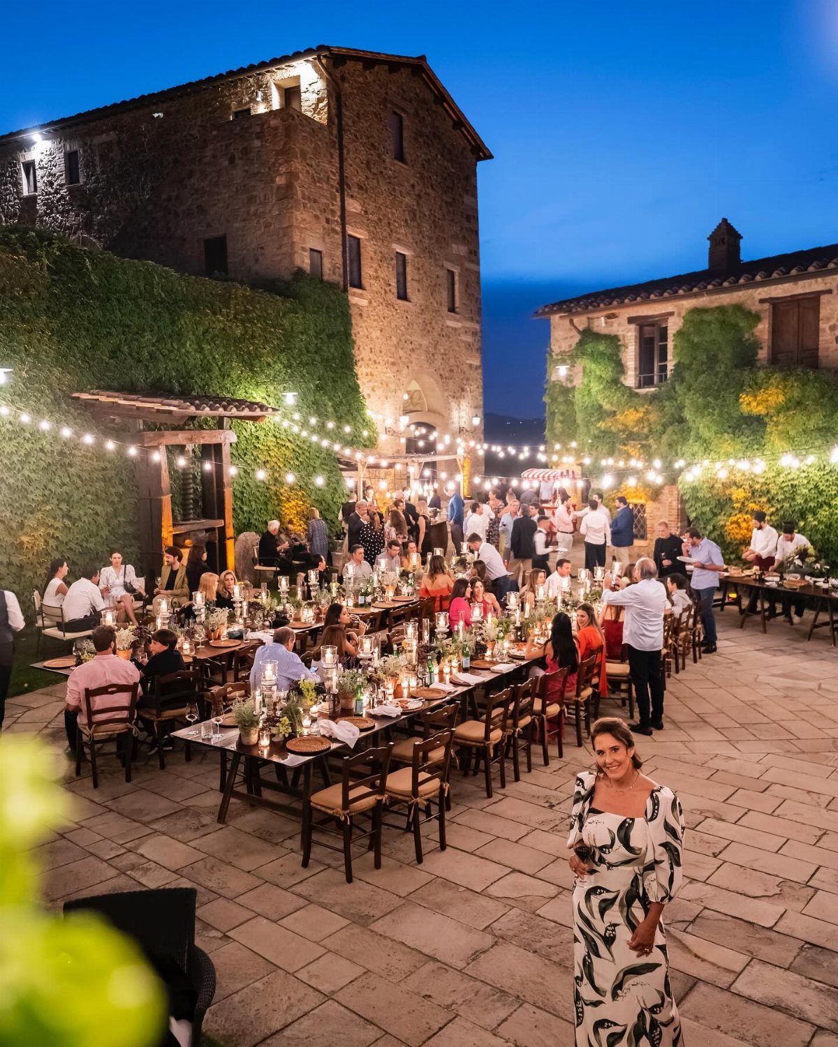 Guests dining at long wooden tables under string lights in a rustic stone courtyard.