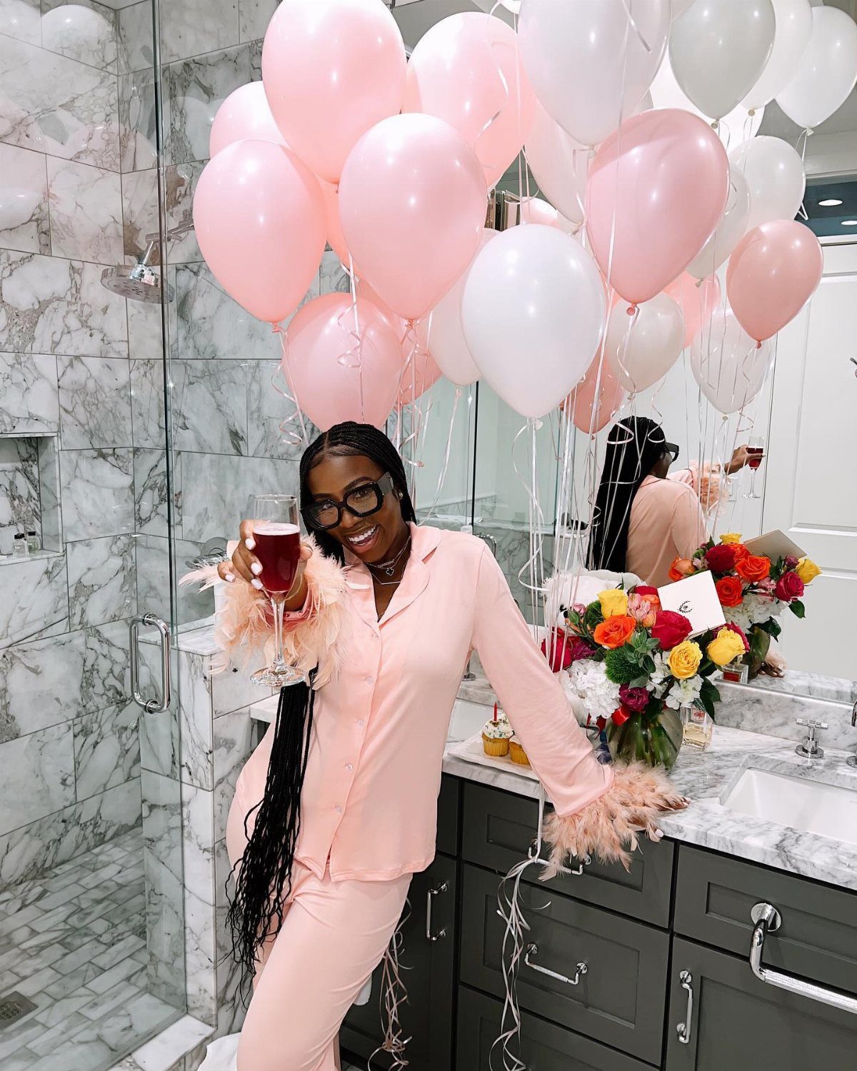 Smiling woman in pink pajamas holding a drink among pink and white balloons.