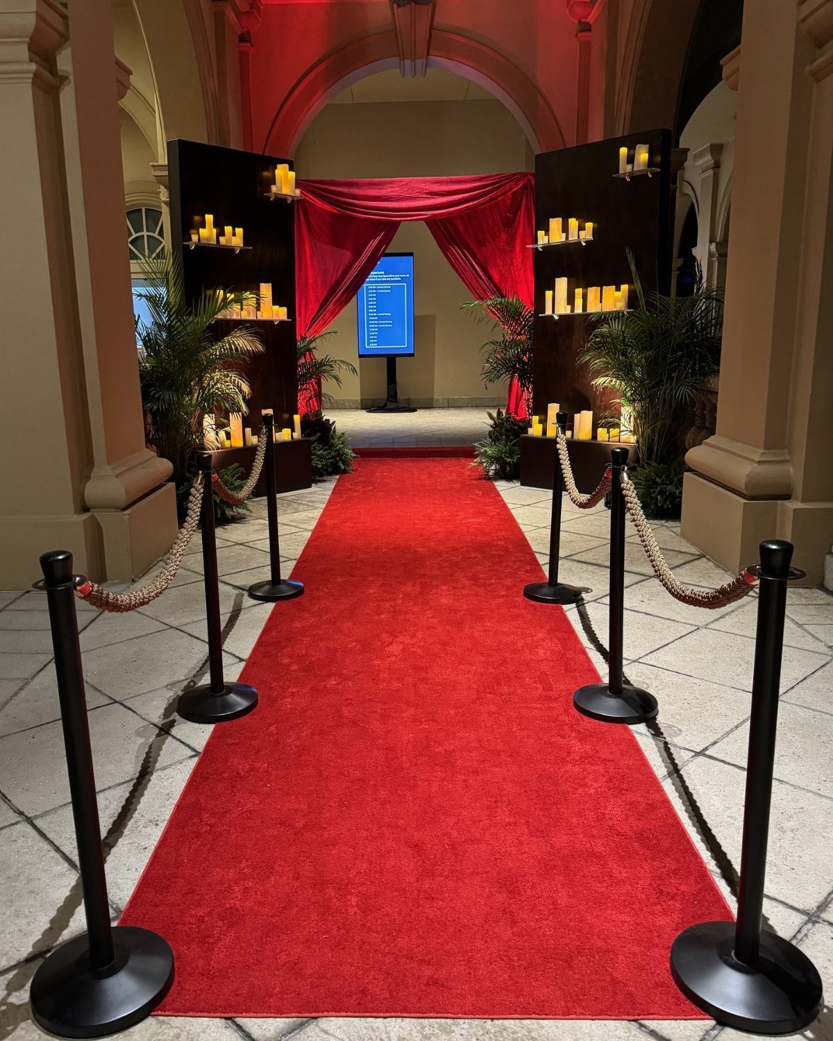 A long red carpet lined with black stanchions and gold ropes leading through an arched entrance.