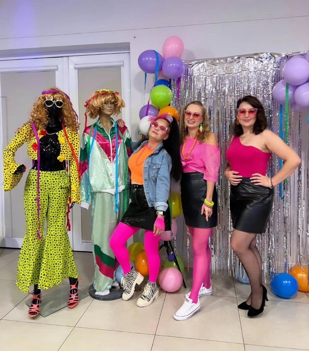 Four women in bright retro clothing posing with mannequins and colorful balloons.