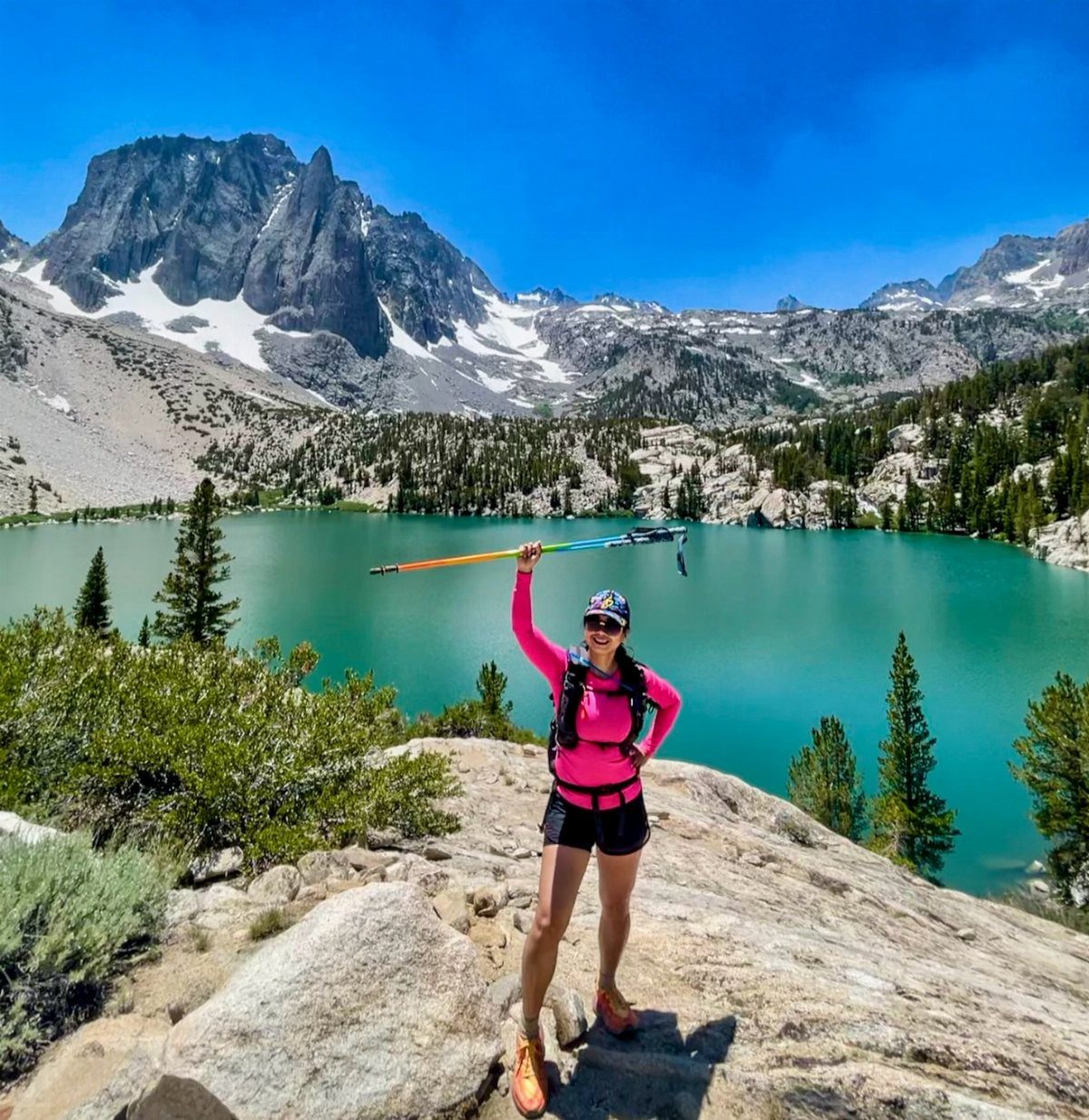 A woman holding a trekking pole stands before a turquoise lake and snowy mountains.
