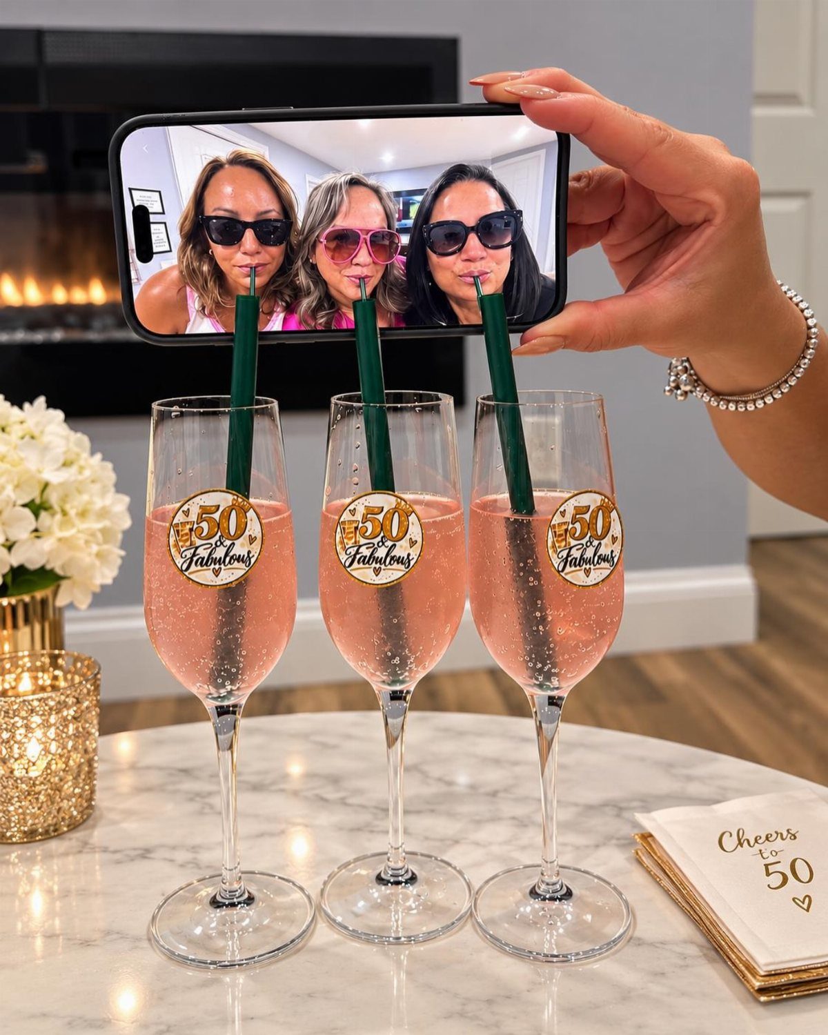 Three champagne flutes with 50 and Fabulous stickers and a smartphone displaying a group selfie.