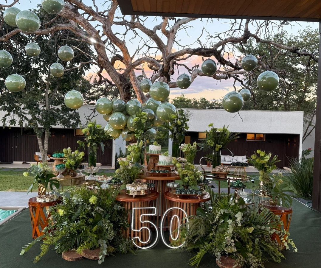 A decorated outdoor dessert table with green balloons, tropical plants, and a neon 50 sign.