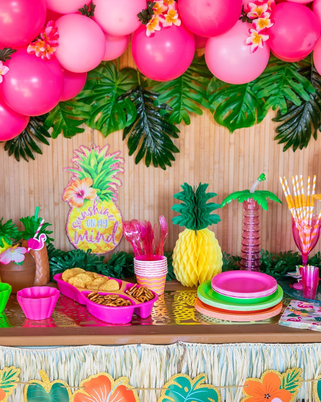 Tropical party table with pink balloons, monstera leaves, pineapple decor, and colorful snacks.