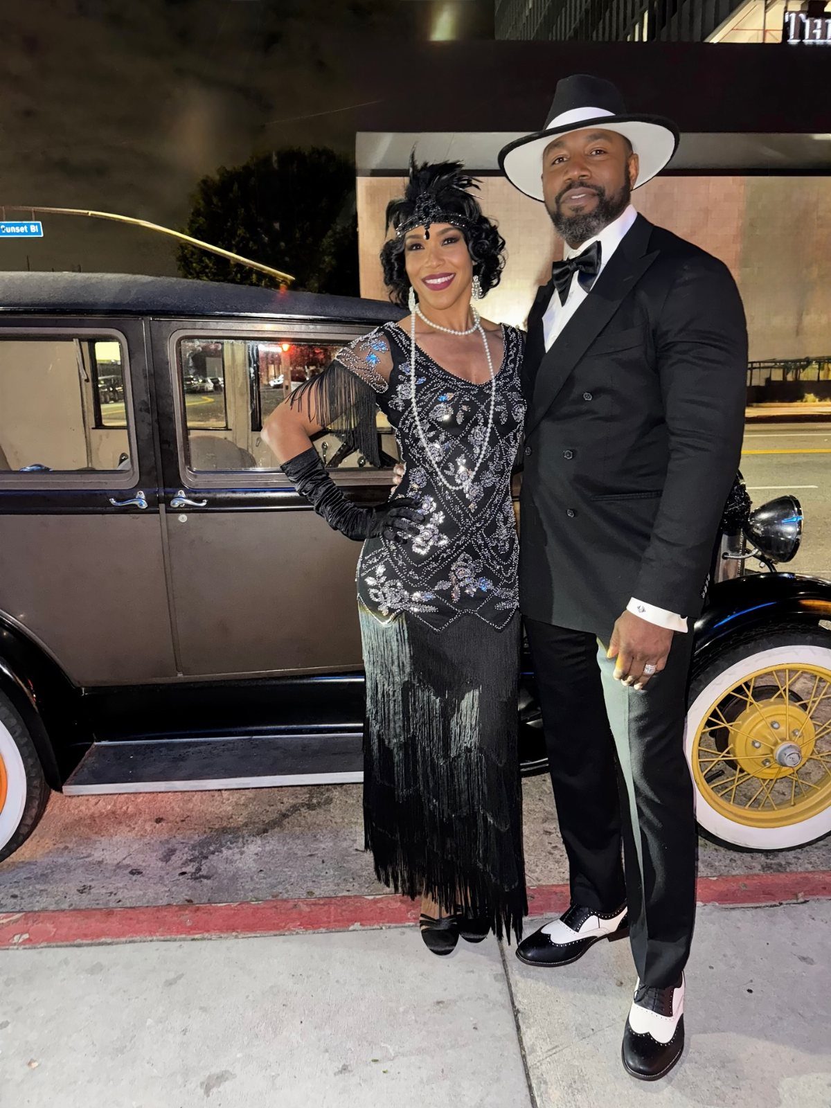 A man and woman in vintage formal attire posing by a classic antique car.