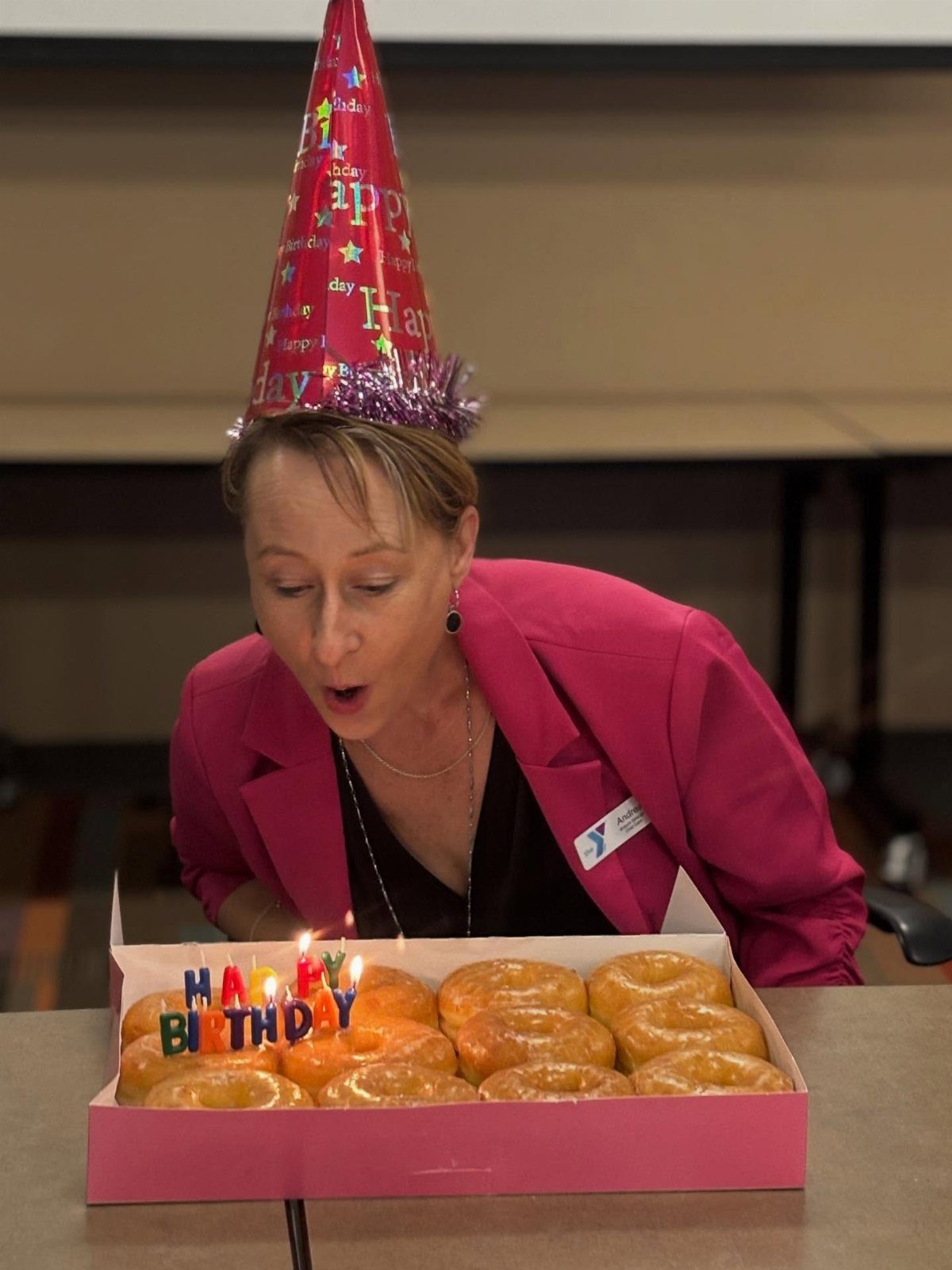 Woman in a pink blazer blowing out candles on a box of glazed donuts.