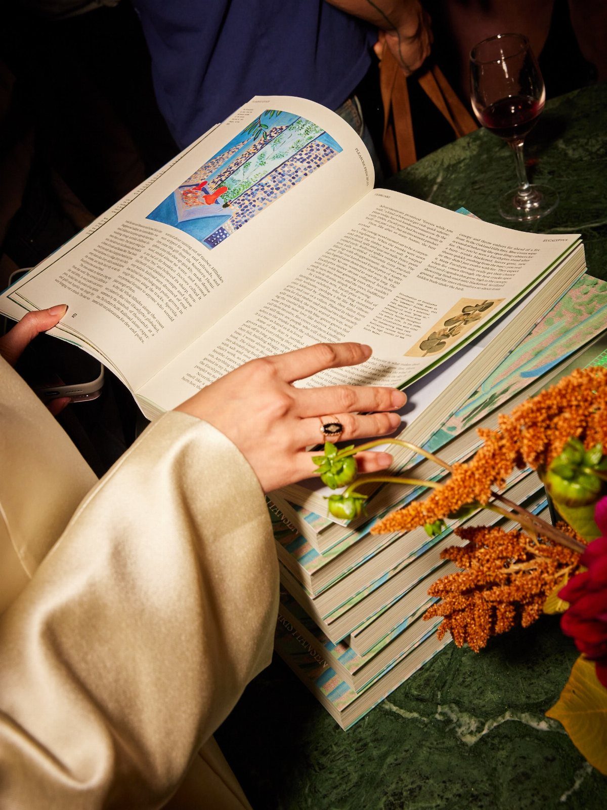 Hands resting on an open book atop a stack of books near red wine.