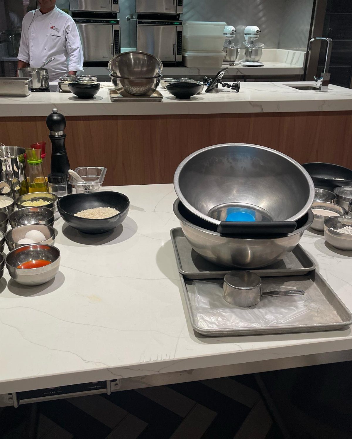 Stainless steel bowls and cooking ingredients arranged on a white kitchen countertop with a chef in the background.