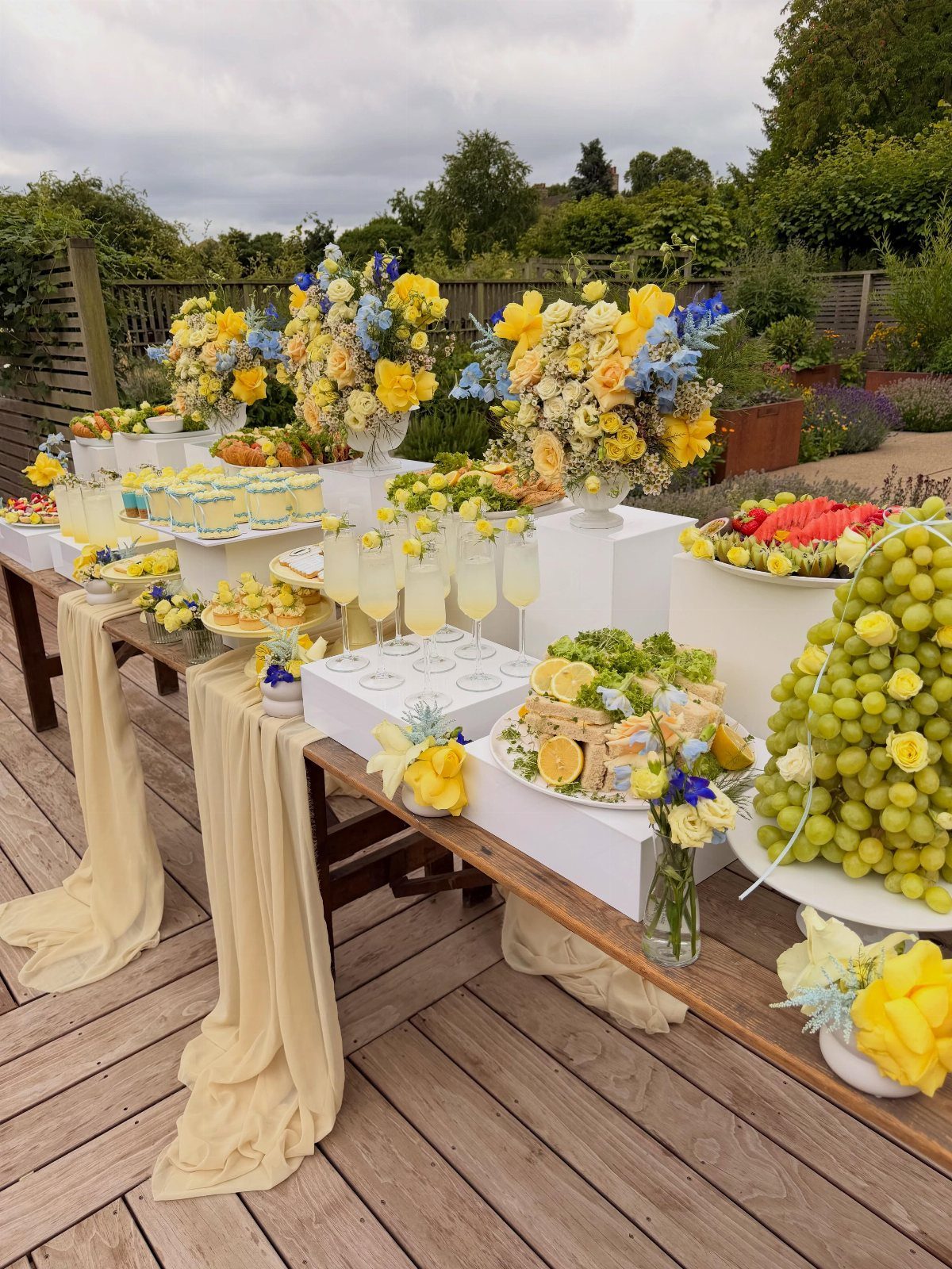 A long outdoor buffet table filled with fruit, cakes, flowers, and drinks on a wooden deck.