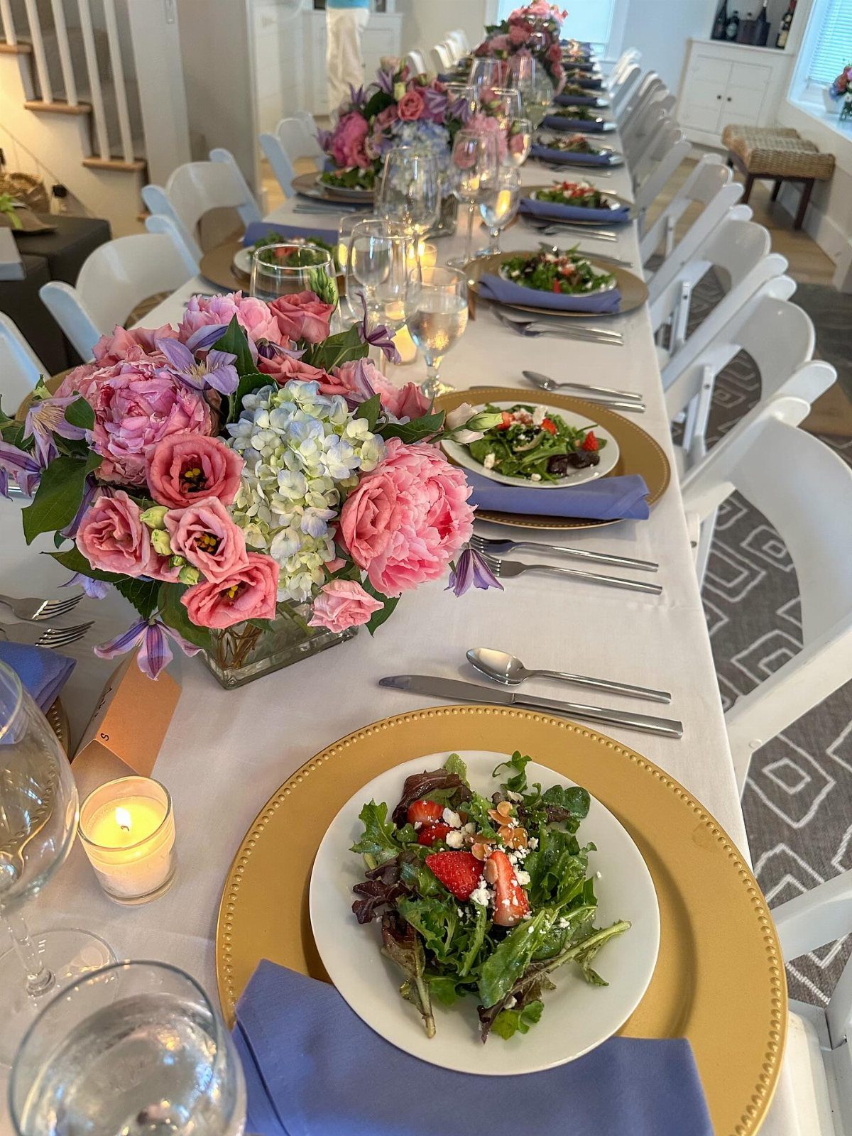 A long, elegantly set dining table featuring floral centerpieces, gold chargers, and fresh salads.