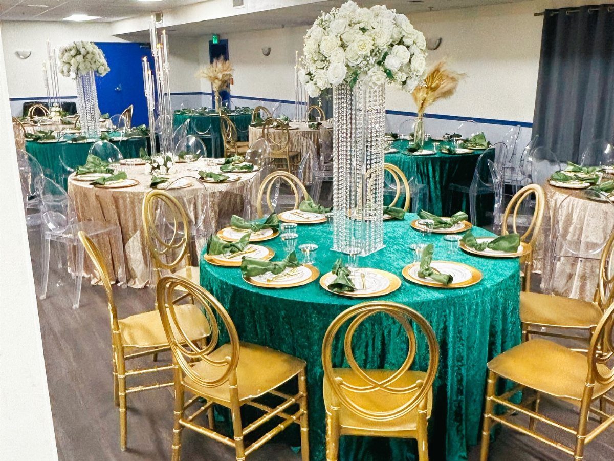 Decorated banquet tables with emerald green cloths, gold chairs, and tall white floral centerpieces in a hall.
