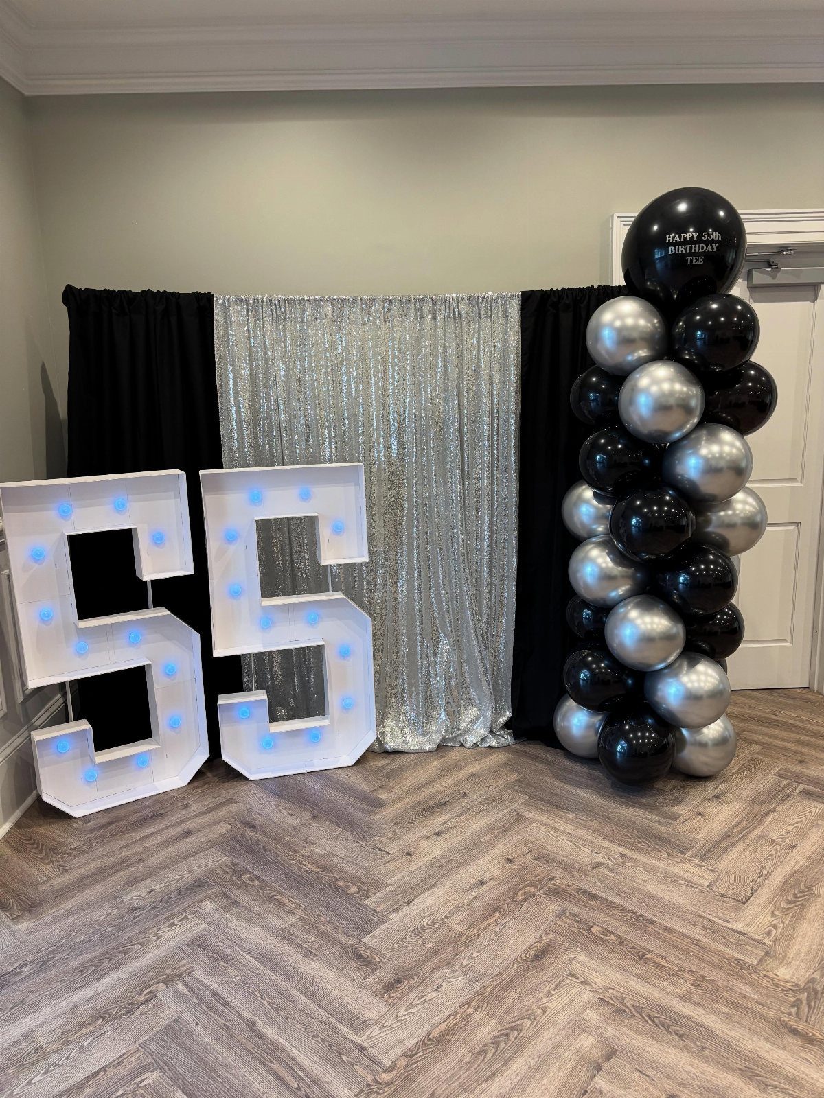 Large illuminated marquee numbers 55 standing before a black and silver sequin backdrop with balloons.