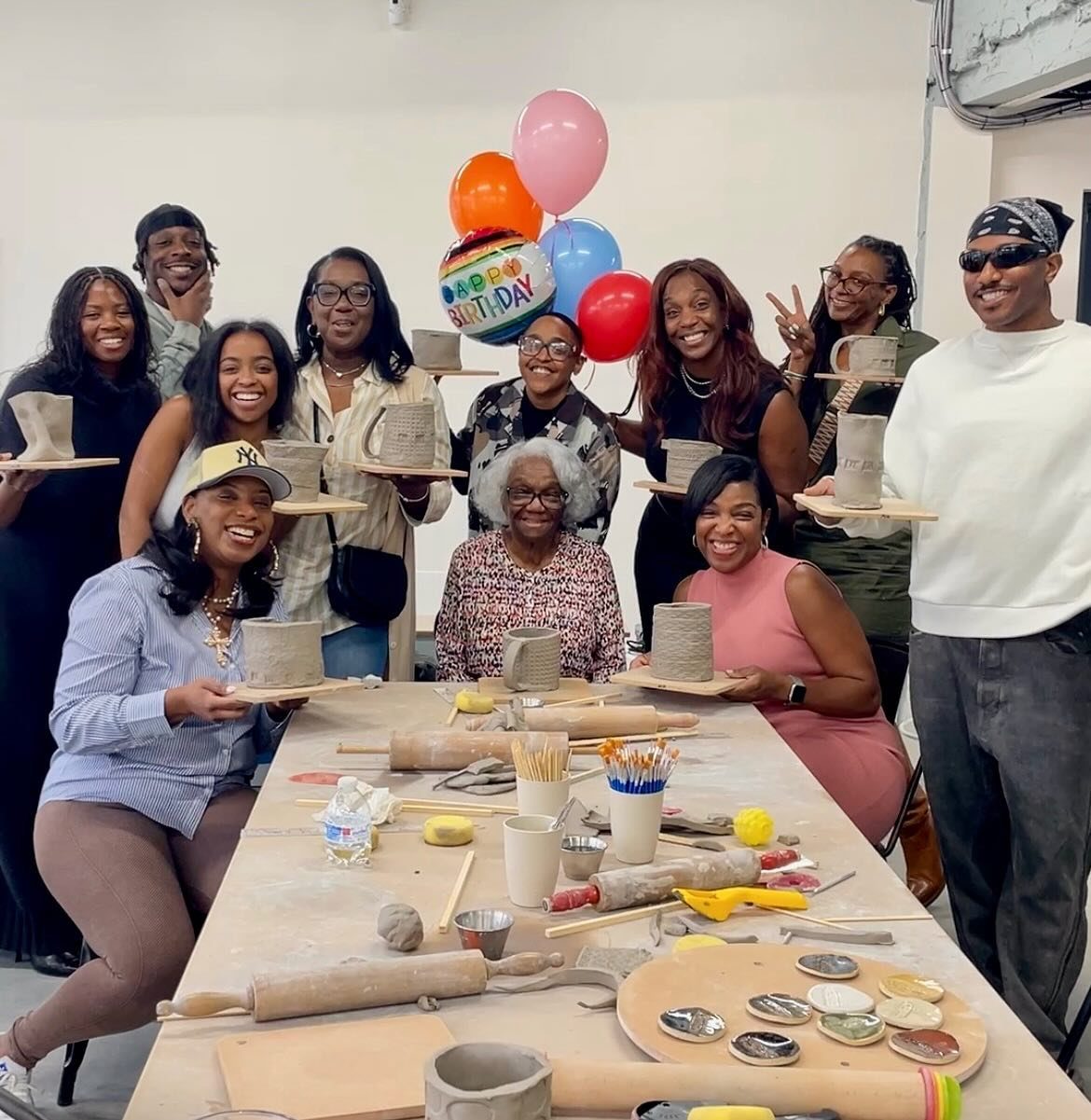 A group of women smiling and holding up handmade pottery pieces around a craft table.