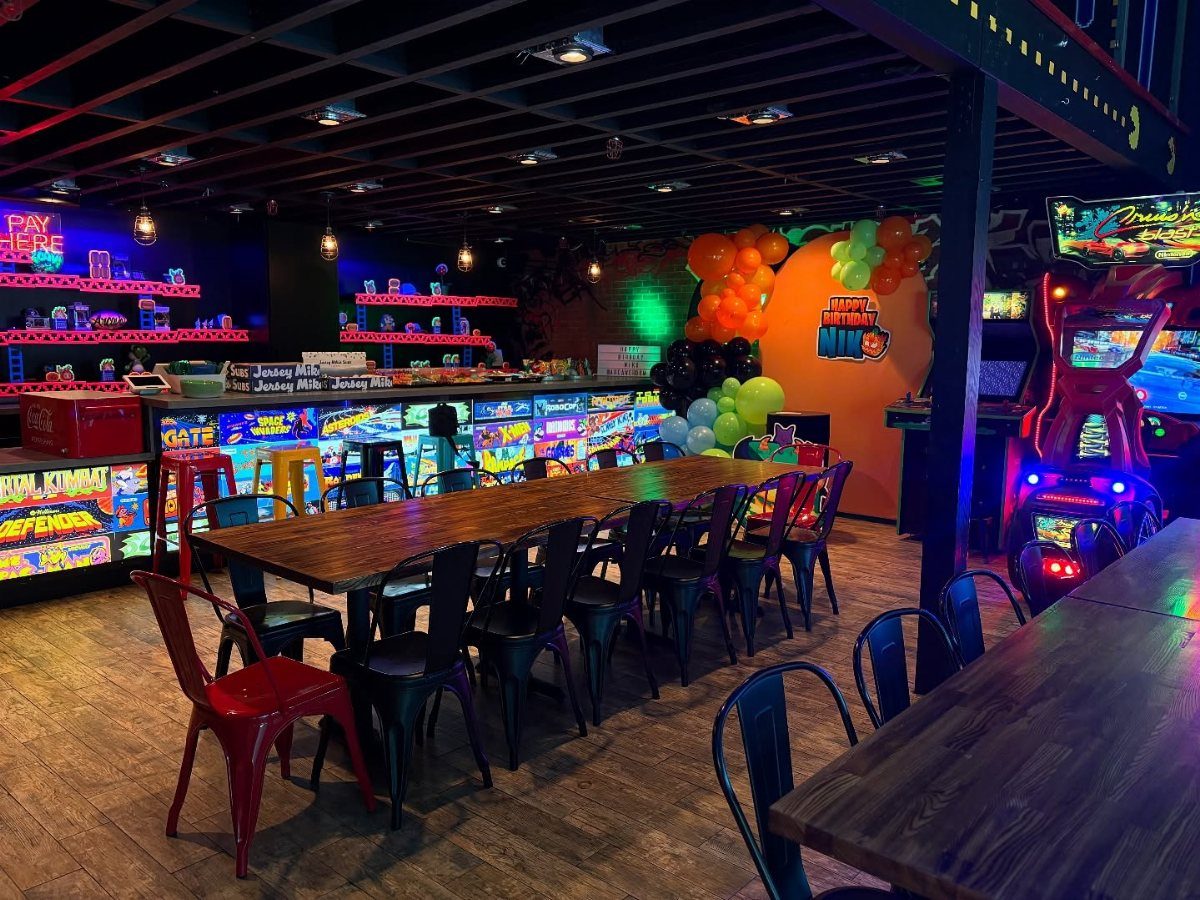 Neon arcade gaming machines and long wooden tables in a brightly lit entertainment venue.