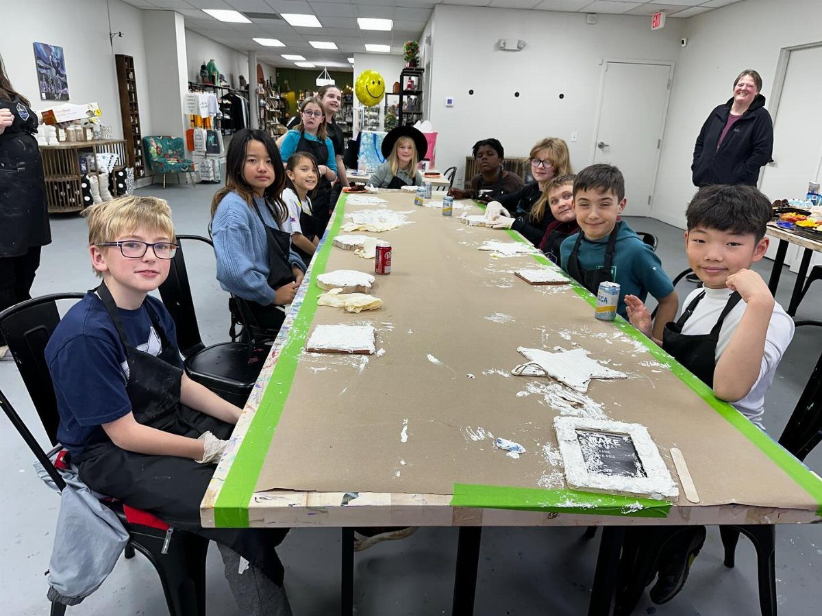 Children wearing aprons sit around a long table covered in craft materials and plaster shapes.