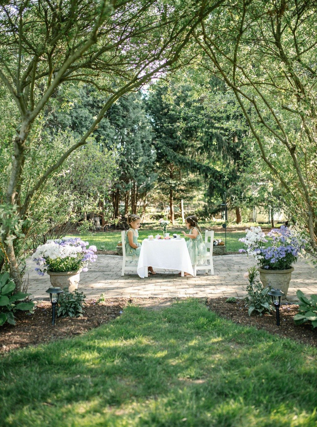 Two young girls sitting at a small white table in a lush green backyard.