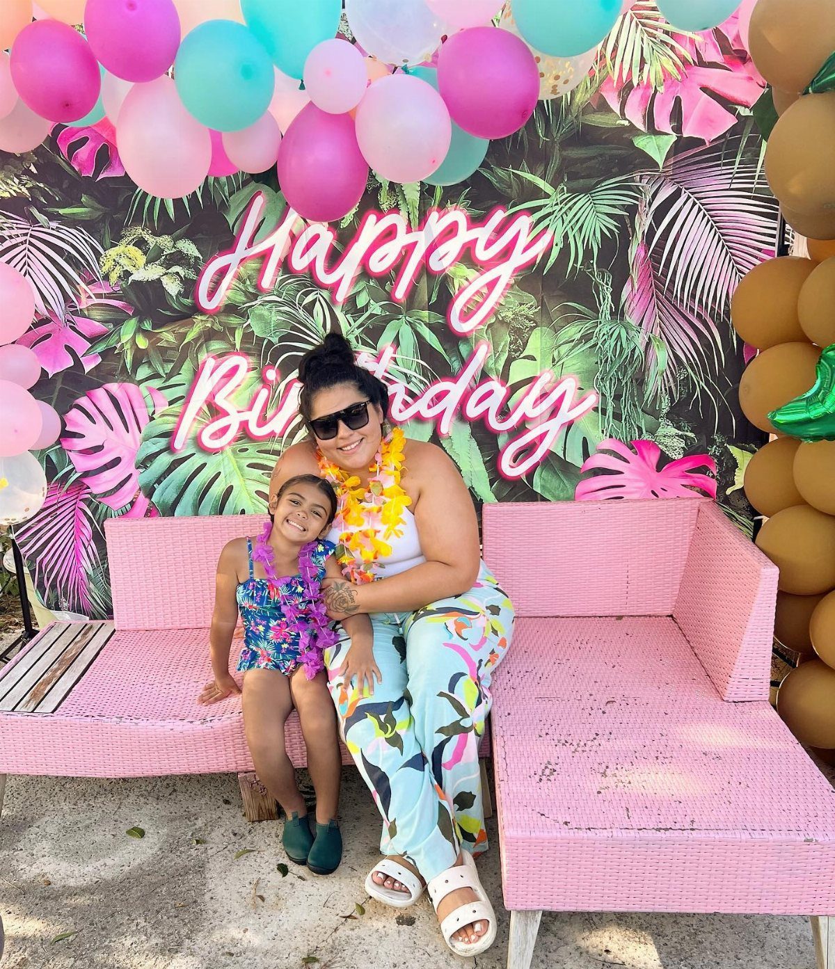 Woman and young girl wearing floral leis sitting on a pink sofa against a tropical backdrop.