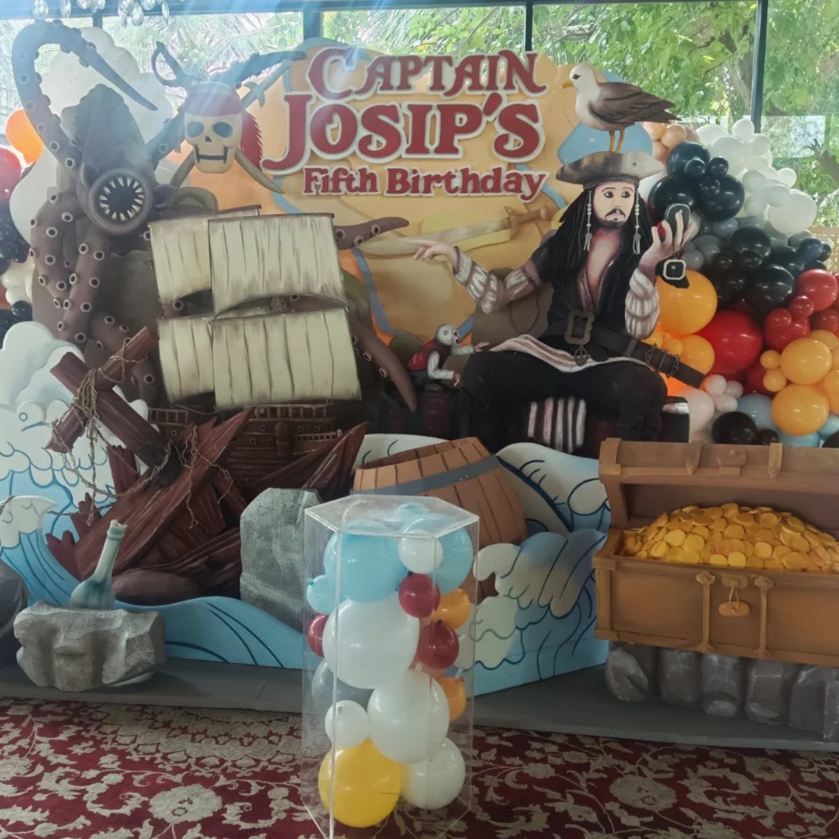 Pirate themed birthday backdrop with a captain cutout, shipwreck, and a treasure chest of gold.
