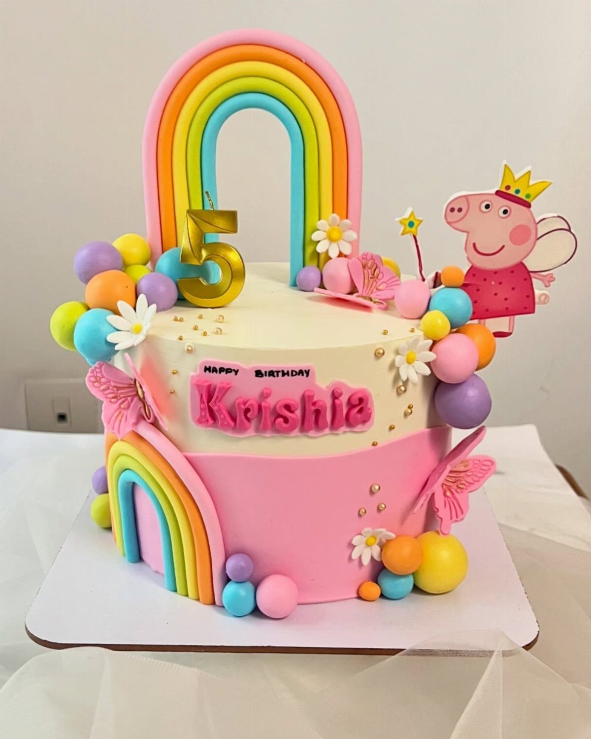 A pink two-tier birthday cake decorated with rainbows, colorful spheres, and a Peppa Pig topper.
