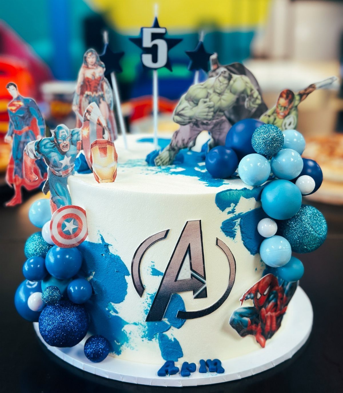 Superhero themed birthday cake with Avengers logo and various character toppers and blue decorations.