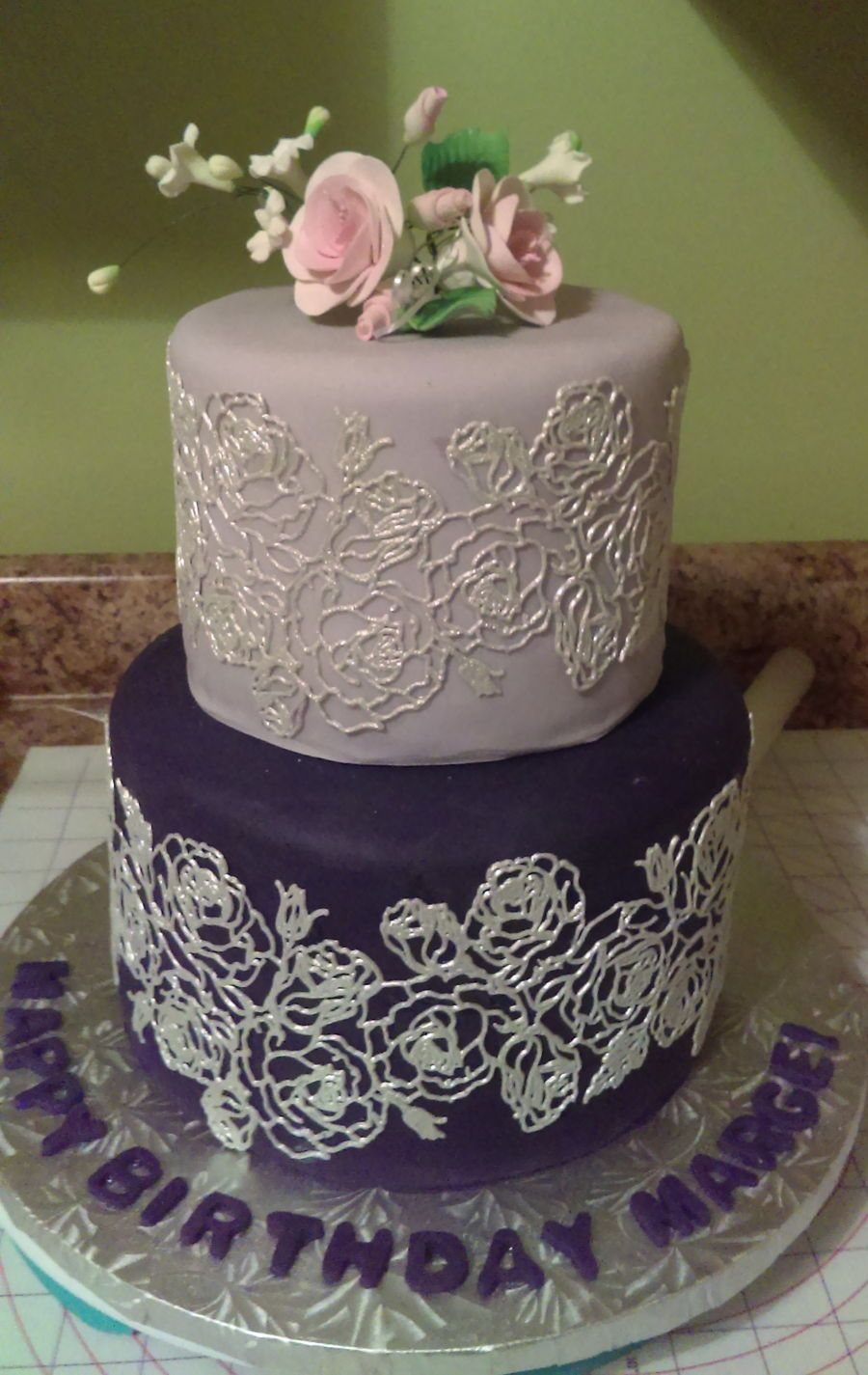 Two-tiered purple cake decorated with intricate silver edible lace and pink sugar flowers.