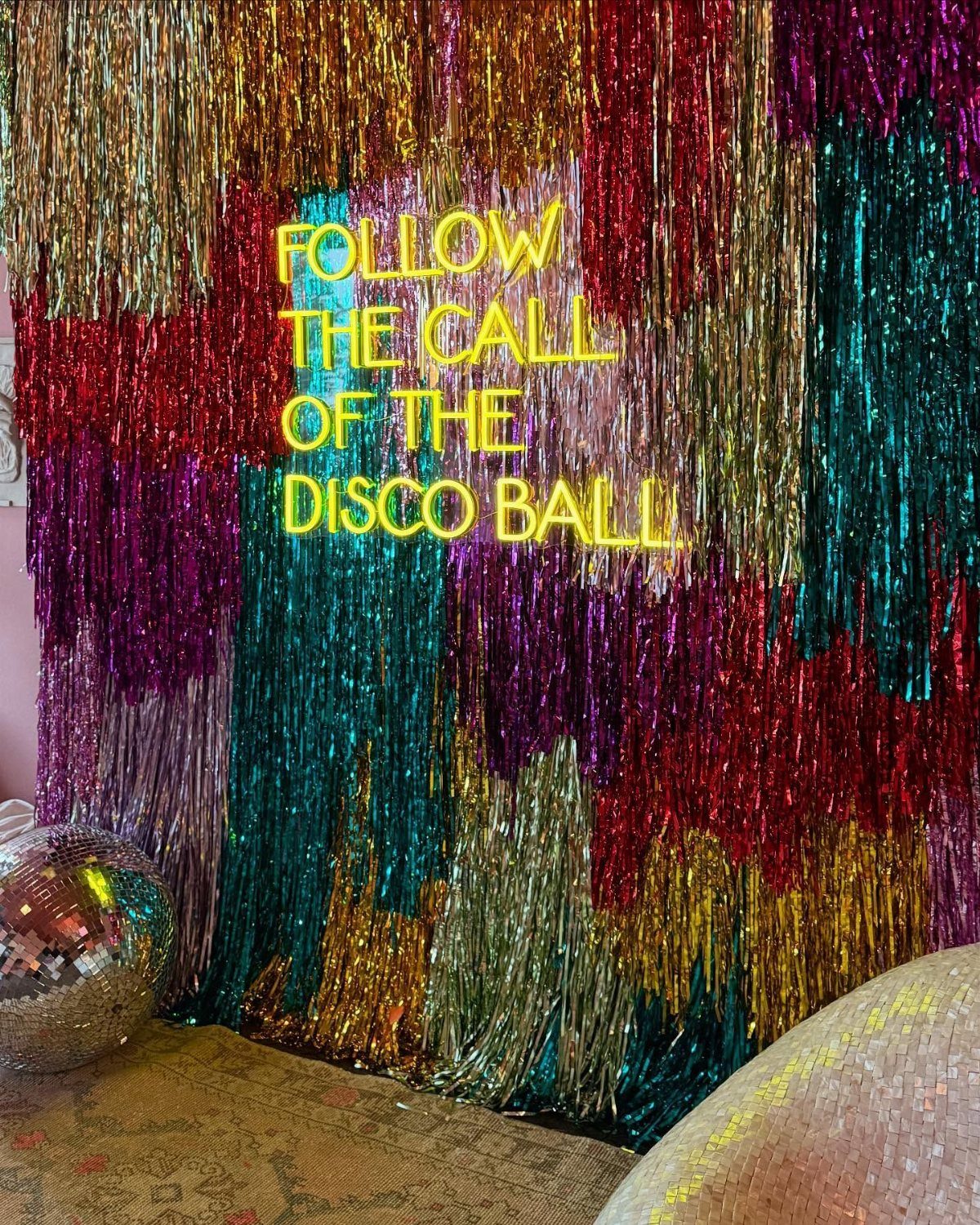 Colorful metallic tinsel backdrop with a yellow neon sign reading Follow the Call of the Disco Ball.
