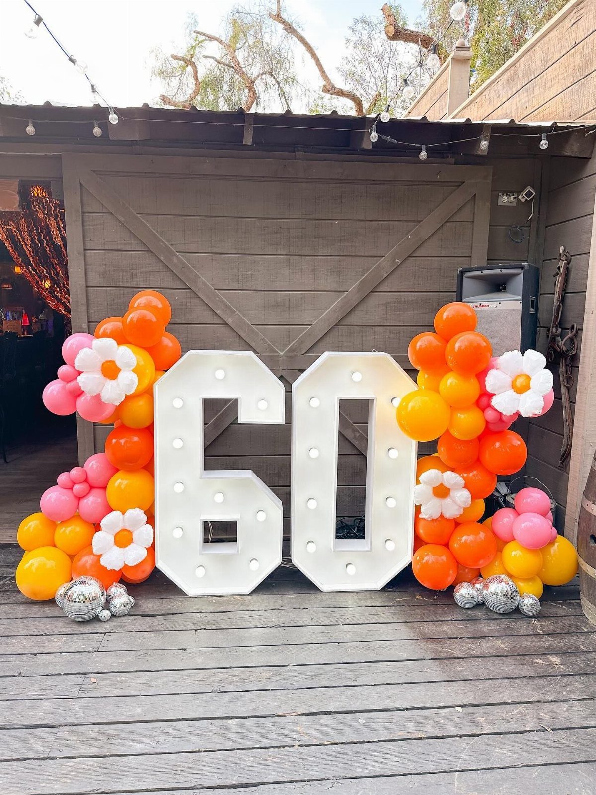 Large illuminated marquee numbers sixty surrounded by orange, pink, and white balloon garlands on a wooden deck.