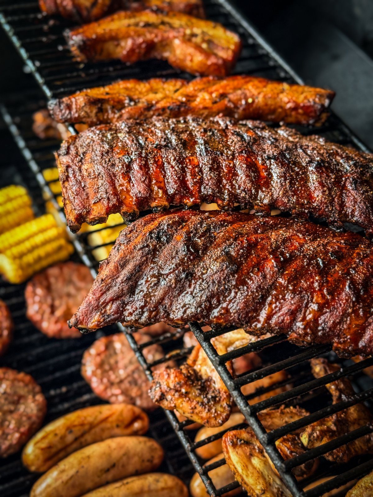 Grilled ribs, chicken wings, sausages, and corn on the cob cooking on a black barbecue grate.