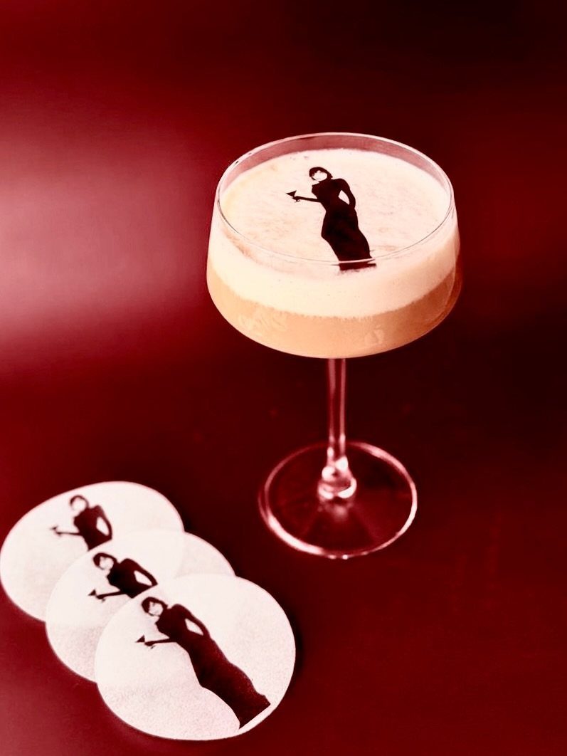 A frothy cocktail in a coupe glass with a woman silhouette on top next to matching coasters.