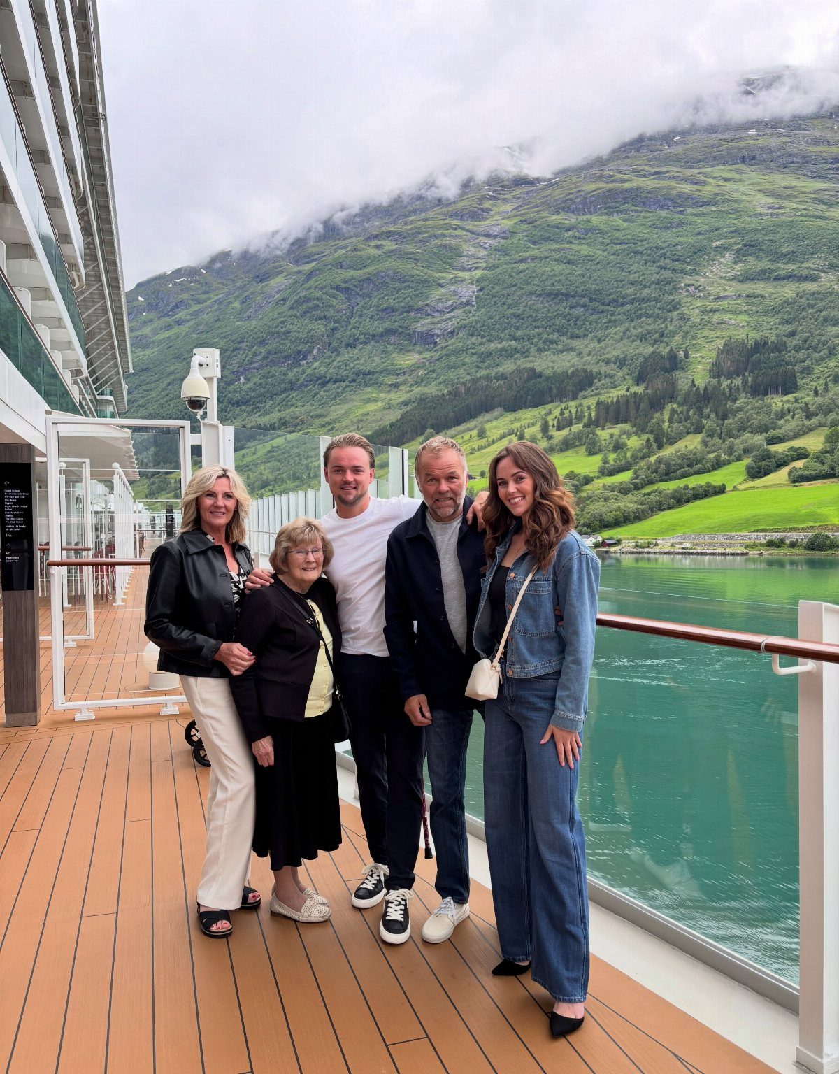 Five people posing together on a cruise ship deck with mountains and water in the background.