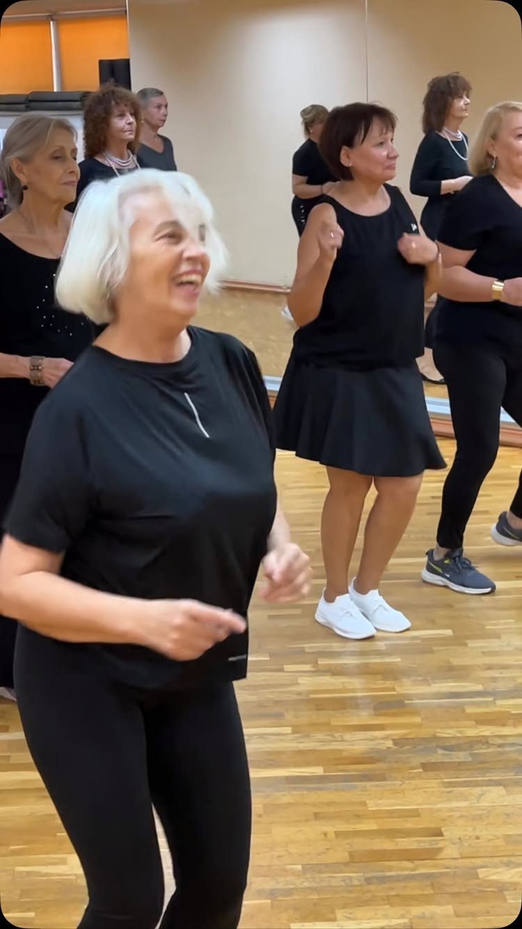 Older women in black activewear dancing together in a bright studio with wooden floors.