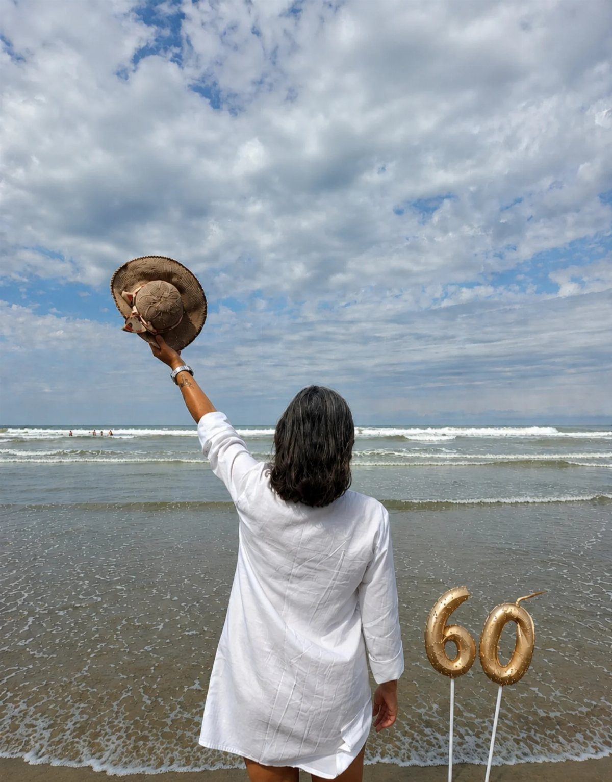 Woman in white shirt holding a straw hat on a beach with 60 balloons.
