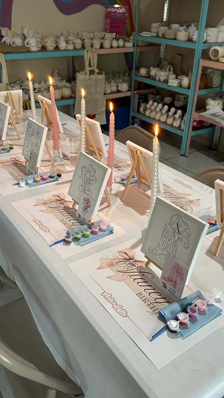 Canvases on small easels with paint palettes and lit candles arranged on a long table.